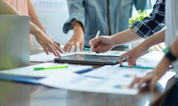 Bild: Projektarbeit an einem Tisch mit Papierunterlagen, Tablet und mehreren Händen.