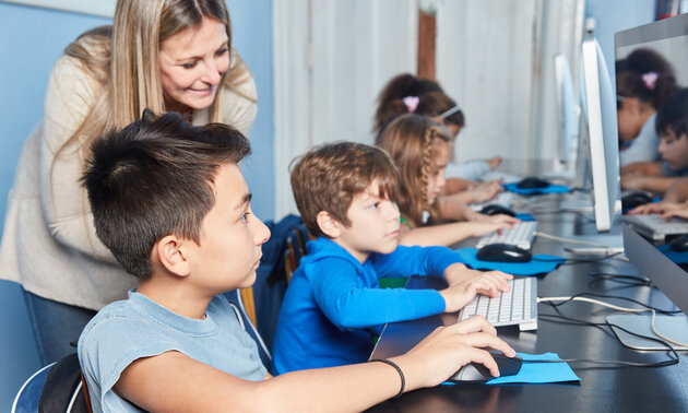 Eine Dame blickt 3 Jungen über die Schulter, die am Computer arbeiten oder spielen