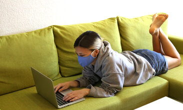 Junges Mädchen liegt auf dem Sofa, arbeitet am Laptop und trägt dabei einen Mund-Nasen-Schutz
