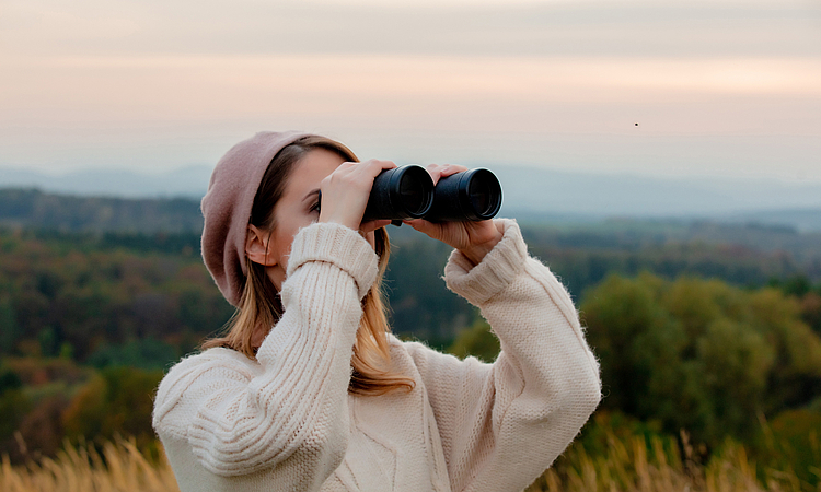 Junge Frau steht auf einem Feld und schaut mit Fernglas in die Ferne