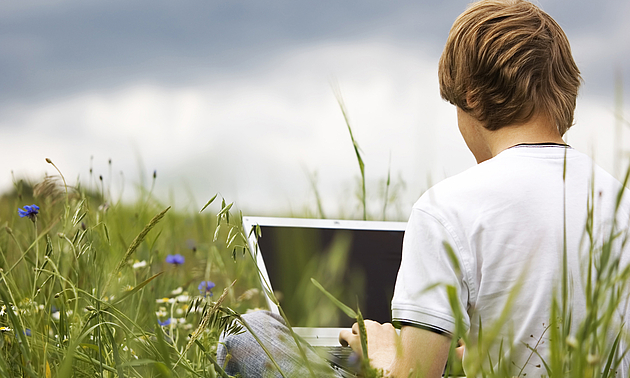 Junger Mann sitzt im Schneidersitz mit augeklapptem Laptop mitten auf einer grünen Wiese