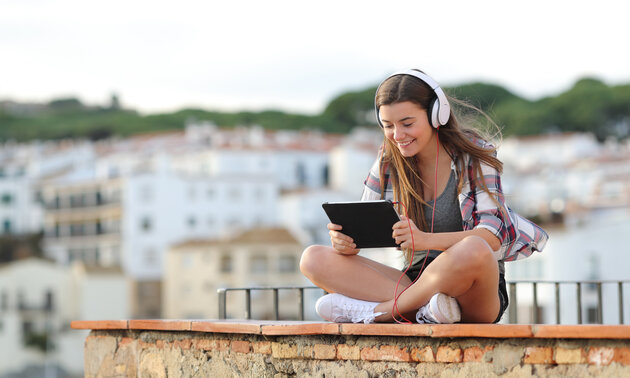 Junge Frau sitzt im Schneidersitz auf einem Dach, schaut lächelnd Inhalte auf dem Tablet und trägt dabei Kopfhörer