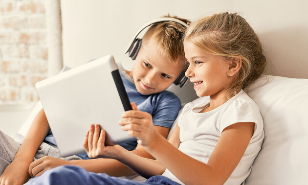 Zwei Kinder sitzen auf einem Sofa und schauen auf ein Tablet